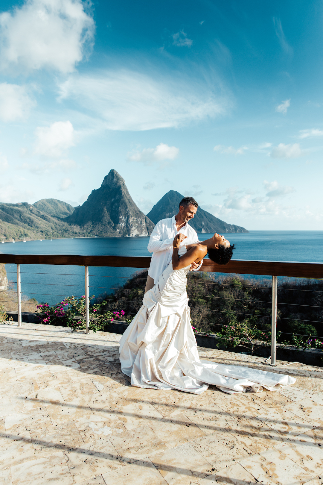 A groom tips his bride back and they are both laughing, against a backdrop of St Lucia's iconic mountains