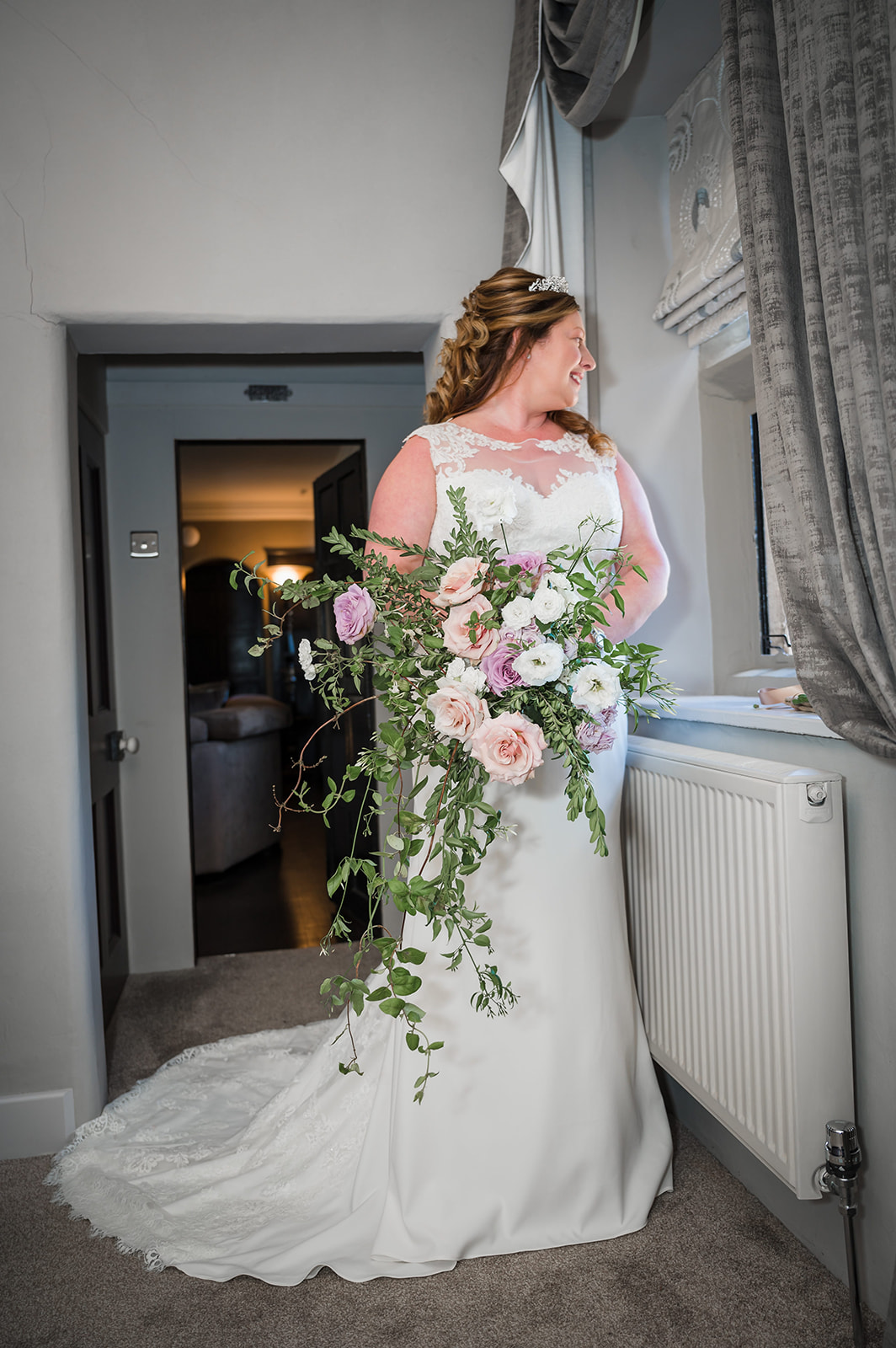 a bride stands in her wedding dress looking out of the window, holding a bouquet of mixed pink and white flowers
